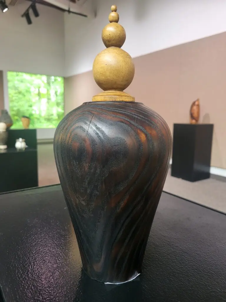 a tall, dark wood urn with visible grain and a lid of stacked spheres carved from light-colored woord.