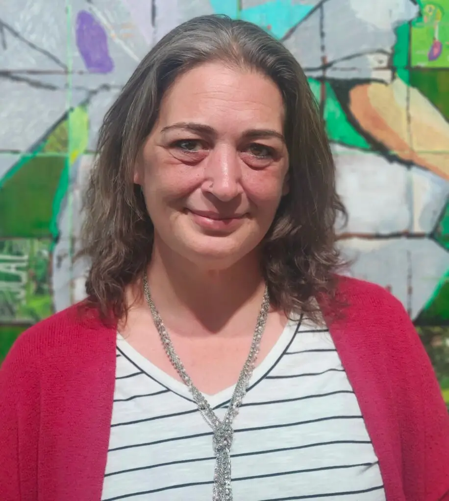 Photo of Theresa Taylor, a white woman with brown hair wearing a red cardigan, white and black striped shirt, and silver necklace standing in front of a multicolored collage.